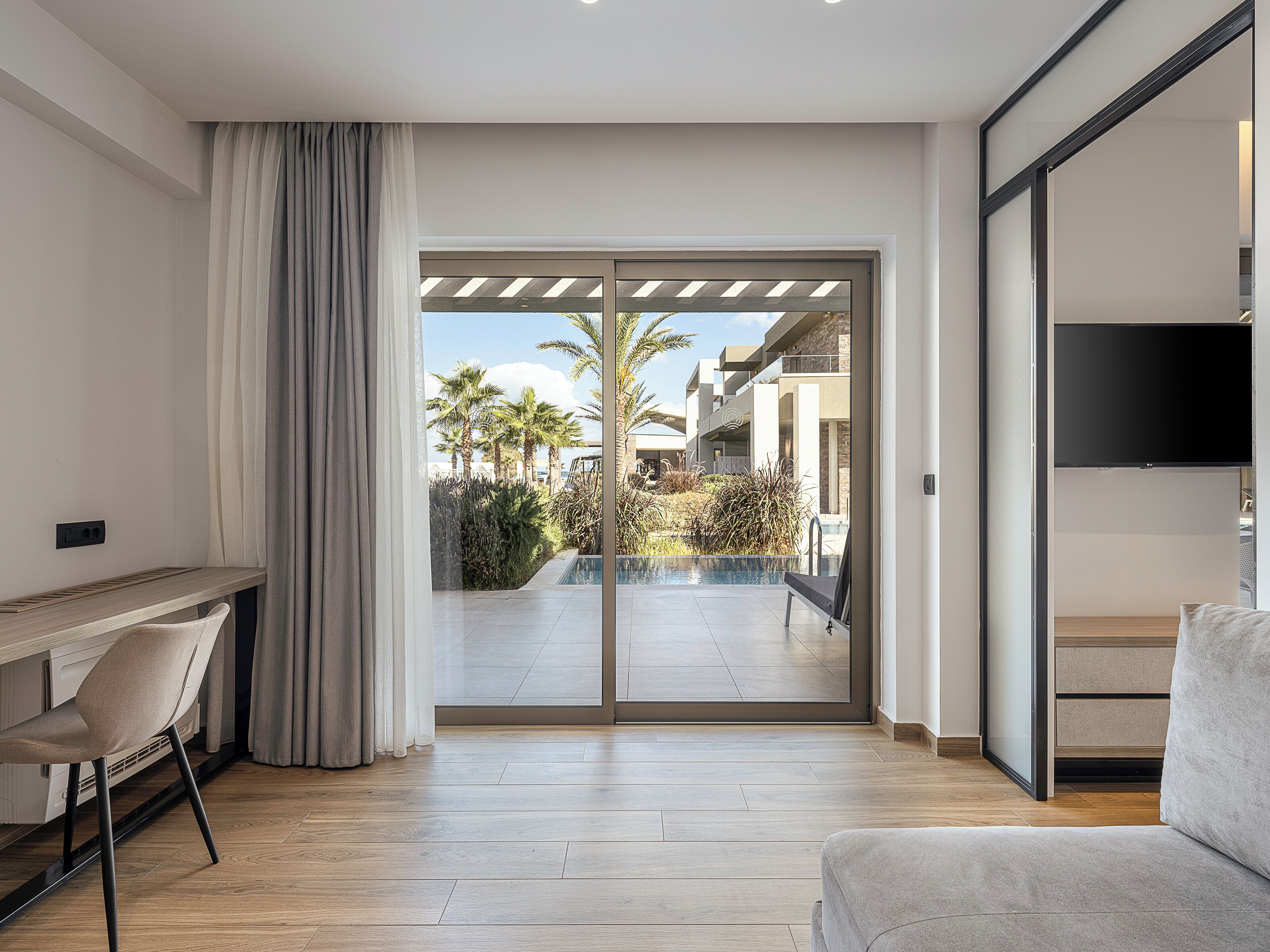 A modern living room with a large sliding glass door leading to a pool area, a flat-screen TV mounted on the wall, and a wooden floor.
