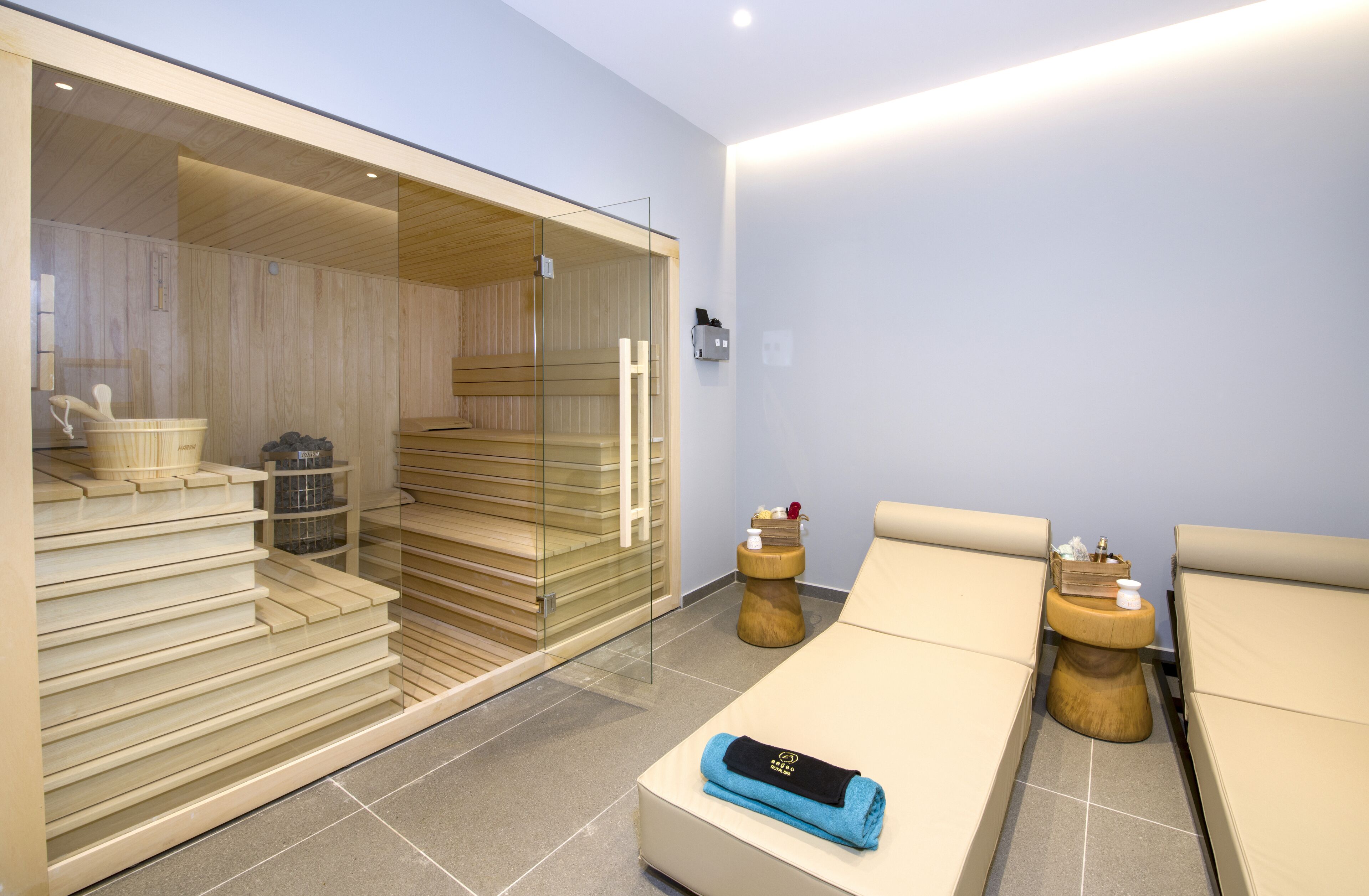 A modern spa room with a wooden sauna, a massage table, and a small wooden stool with a book and a candle.