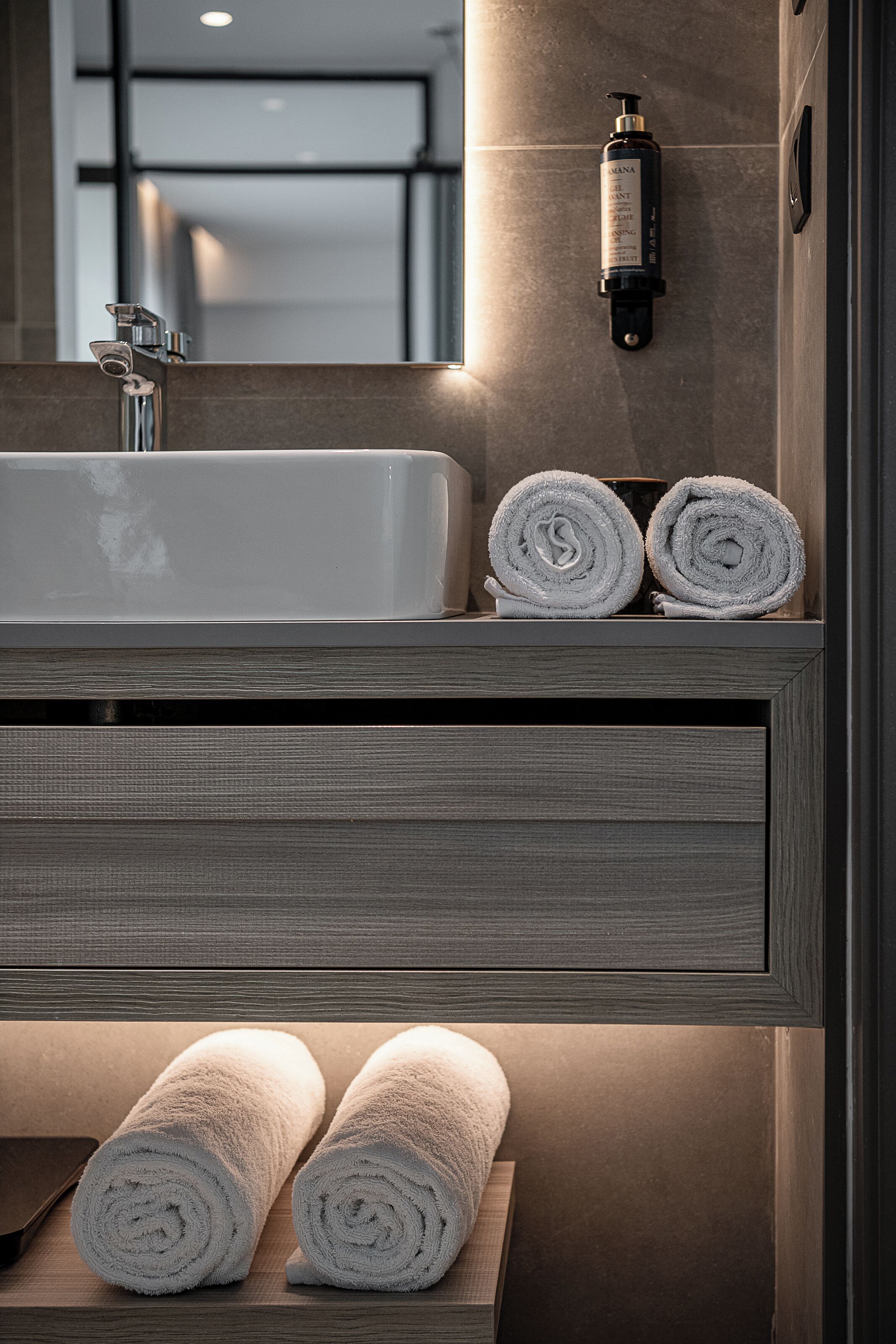 Un baño con un lavabo grande, dos toallas enrolladas en un estante y una botella de champú en la pared.
