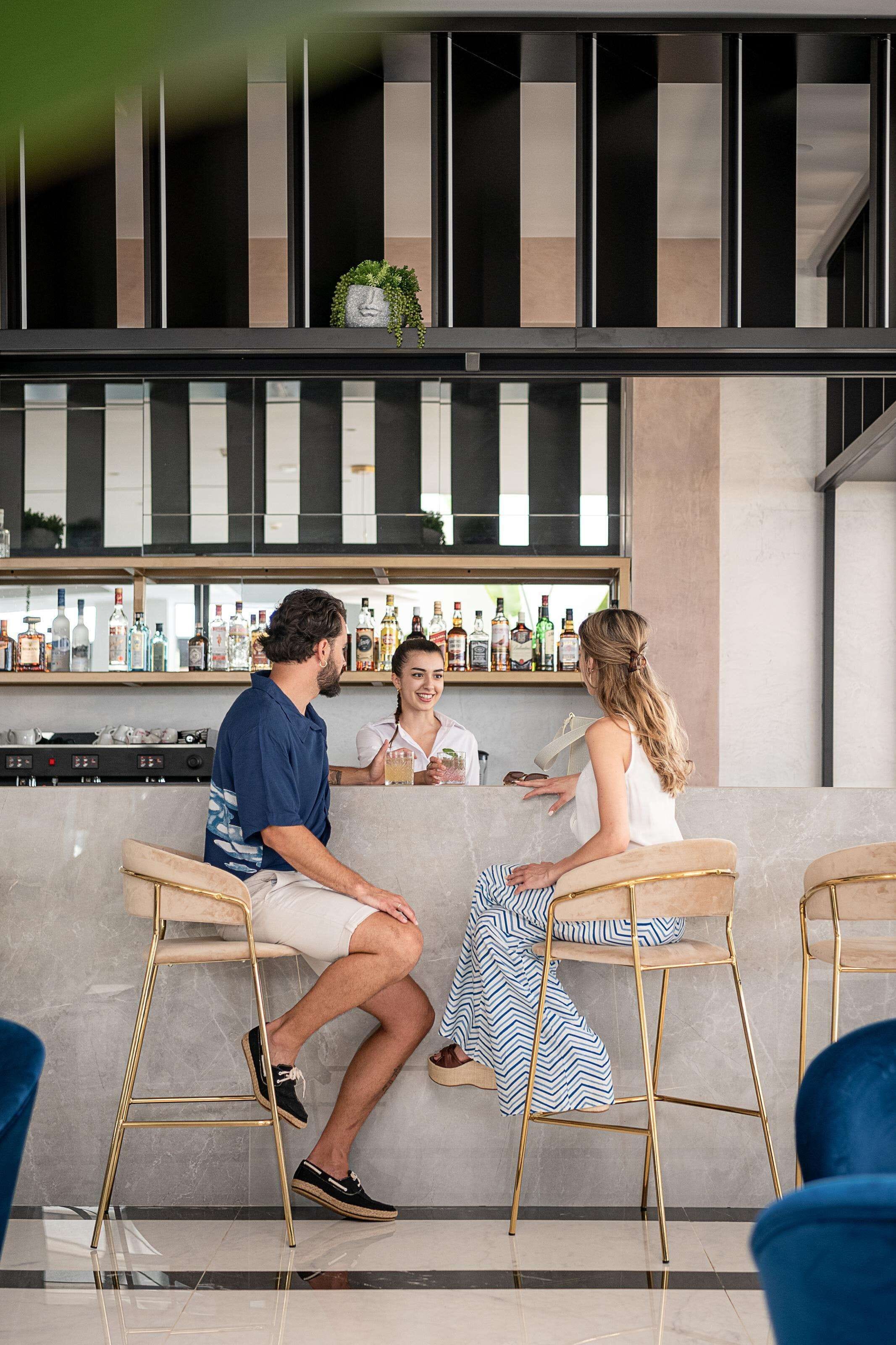 A bar area with a counter displaying various bottles, two bar stools, and a seating area with a person seated.