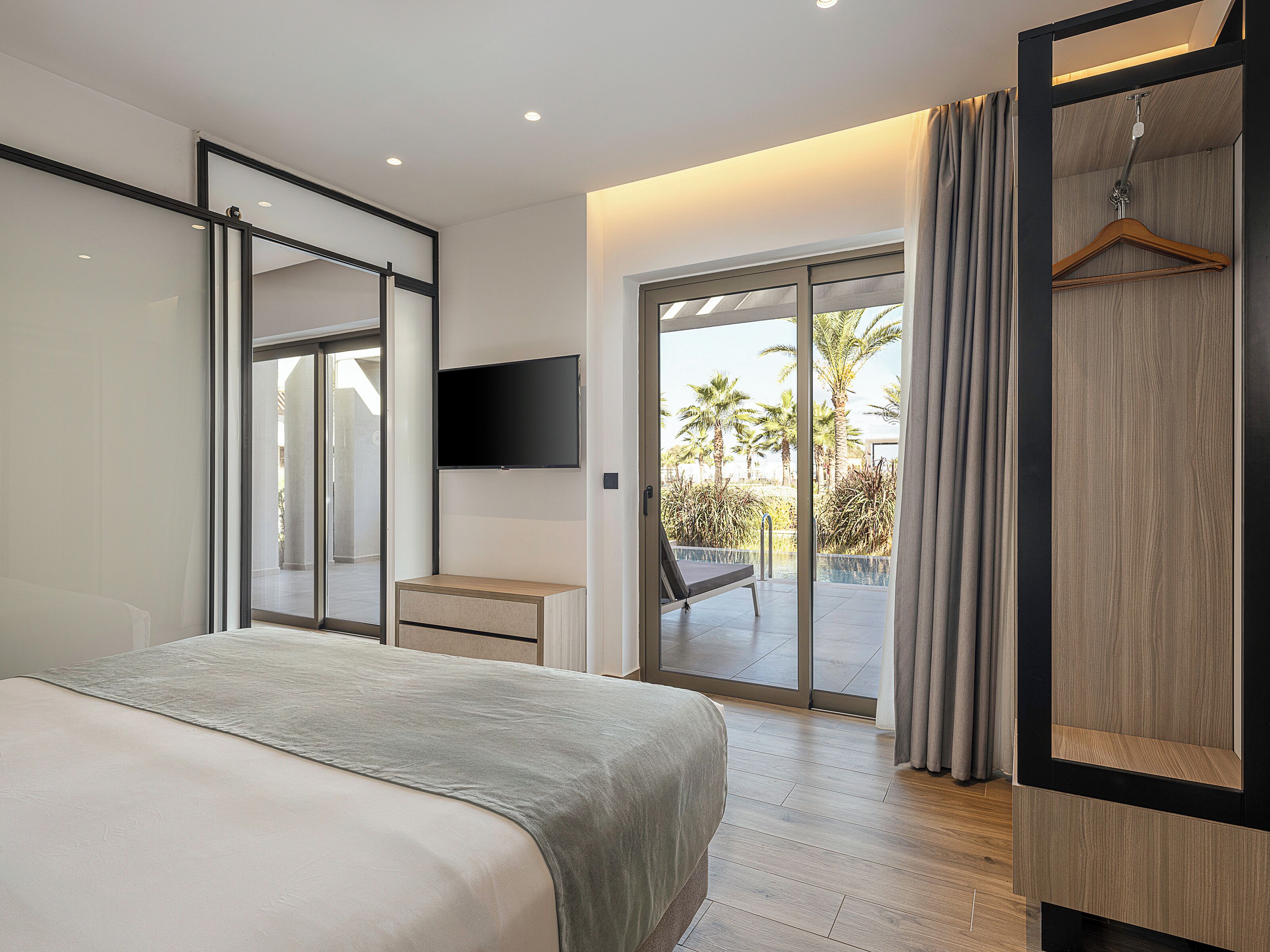 Ein modernes Schlafzimmer mit Bett, Schreibtisch, Fernseher und einer Schiebetür zu einem Balkon mit Pool und Palmen.