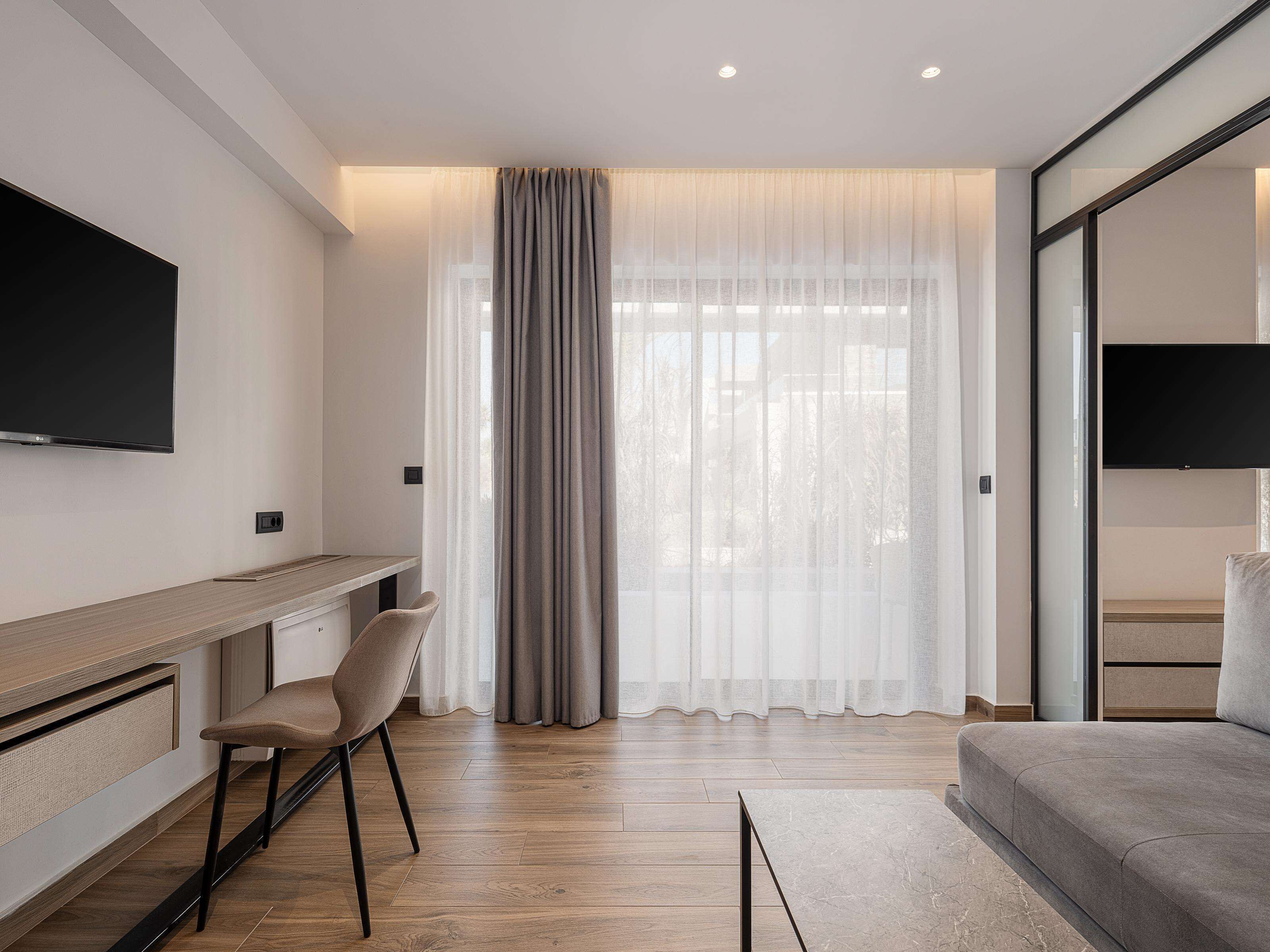 A modern hotel room with a flat-screen TV, a wooden desk, a comfortable chair, a sofa, and a large window with sheer curtains.