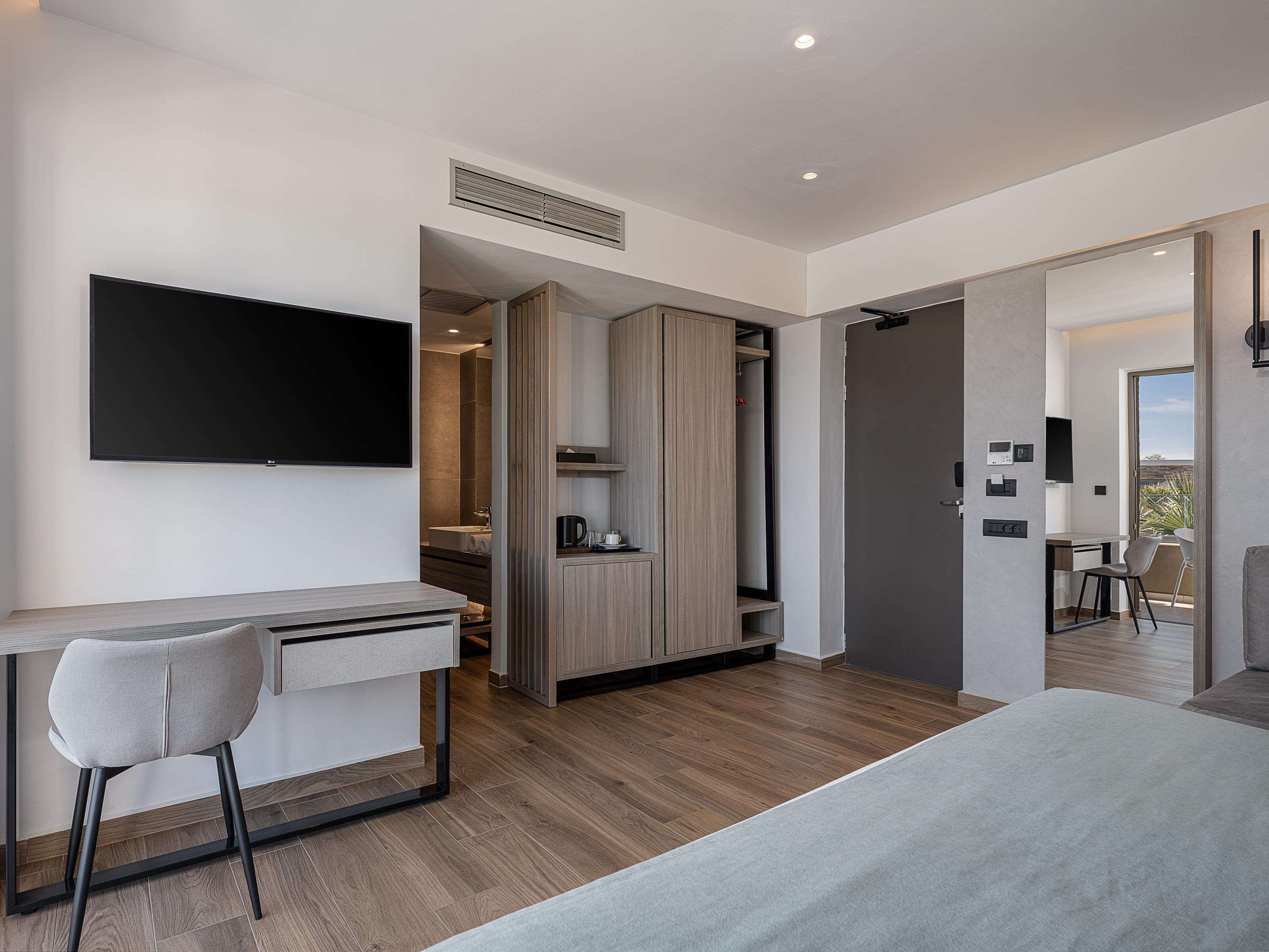 A modern hotel room with a bed, a desk, and a chair. There is a TV mounted on the wall, a wardrobe, and a bathroom visible through an open door.