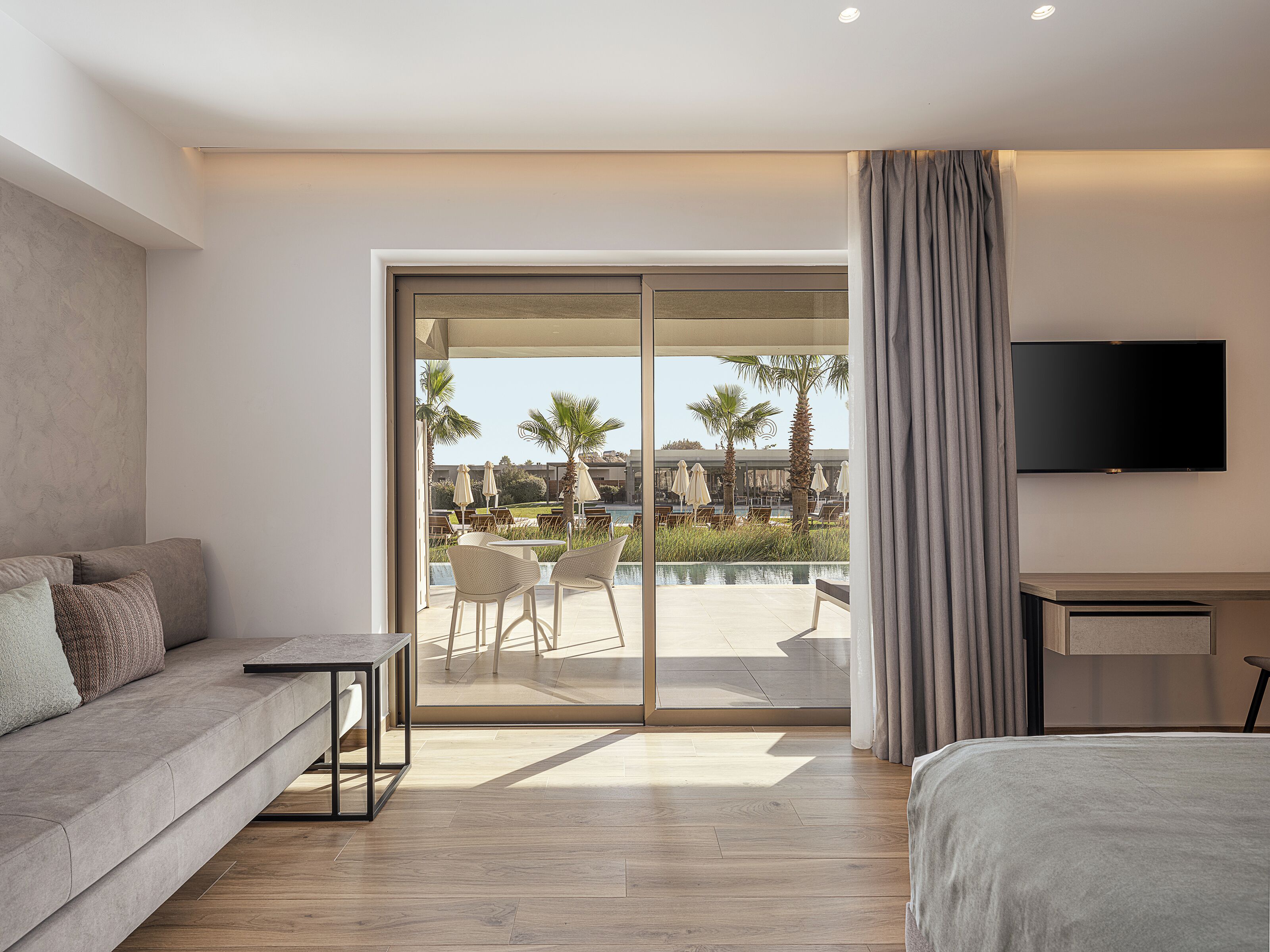 Una habitación de hotel moderna con sofá, cama, televisor y puerta corrediza de vidrio que da a una zona de piscina con mobiliario exterior.