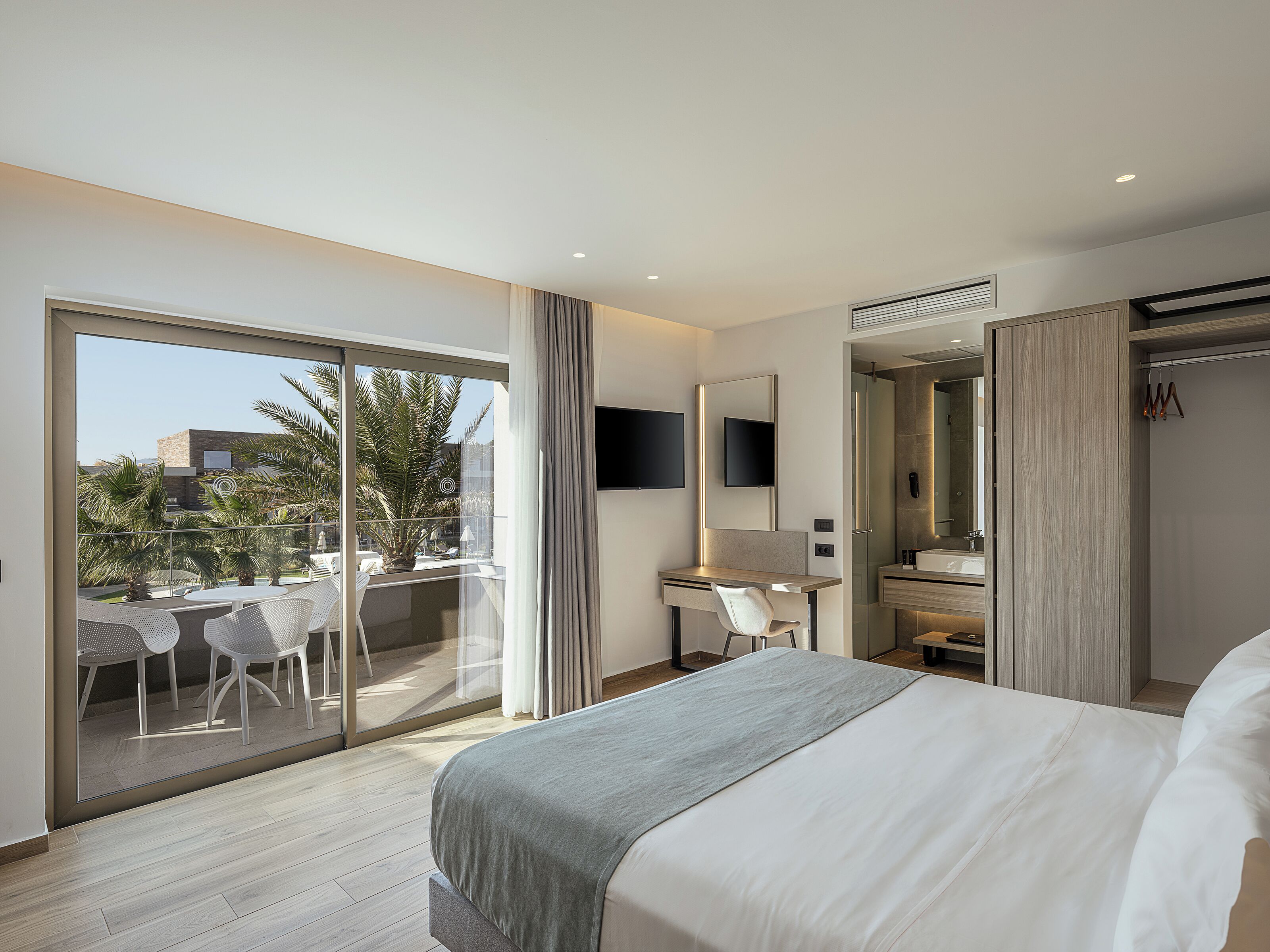 A modern hotel room with a large bed, a desk with a chair, a TV, and a balcony with outdoor seating.