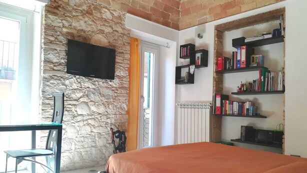 A bedroom with a stone wall, a bed with an orange bedspread, a TV mounted on the wall, and a bookshelf filled with books. - Colle di Minerva Entire Apartment (Chieti)