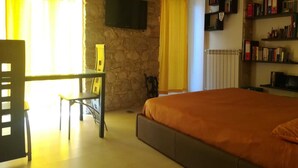 A bedroom with a stone wall, a bed with an orange bedspread, a desk with a chair, a TV mounted on the wall, and a bookshelf with books. - Colle di Minerva Entire Apartment (Chieti)
