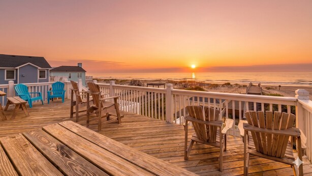 A wooden deck with chairs and a view of the sunset over the ocean. - Oceanfront luxury in this spacious 10-bedroom home in South Nags Head. (South Nags Head)