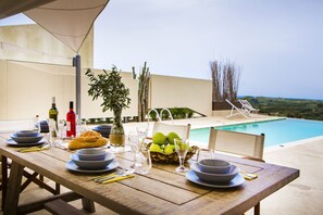 A wooden dining table set with plates, bowls, cutlery, and a basket of green apples, overlooking a swimming pool and a scenic landscape.