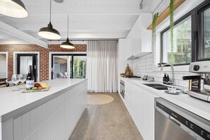 A modern kitchen with white cabinetry, a large island, hanging plants, and a view to the outside. - Sorrento Beach House (Sorrento)