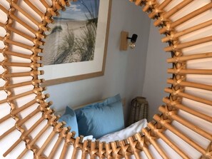 A room with a circular woven headboard, a framed picture of a beach scene, a blue pillow, and a small lamp on the wall.
