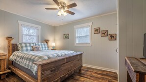 Un dormitorio con una cama de madera, dos ventanas, un ventilador de techo y obras de arte en la pared.