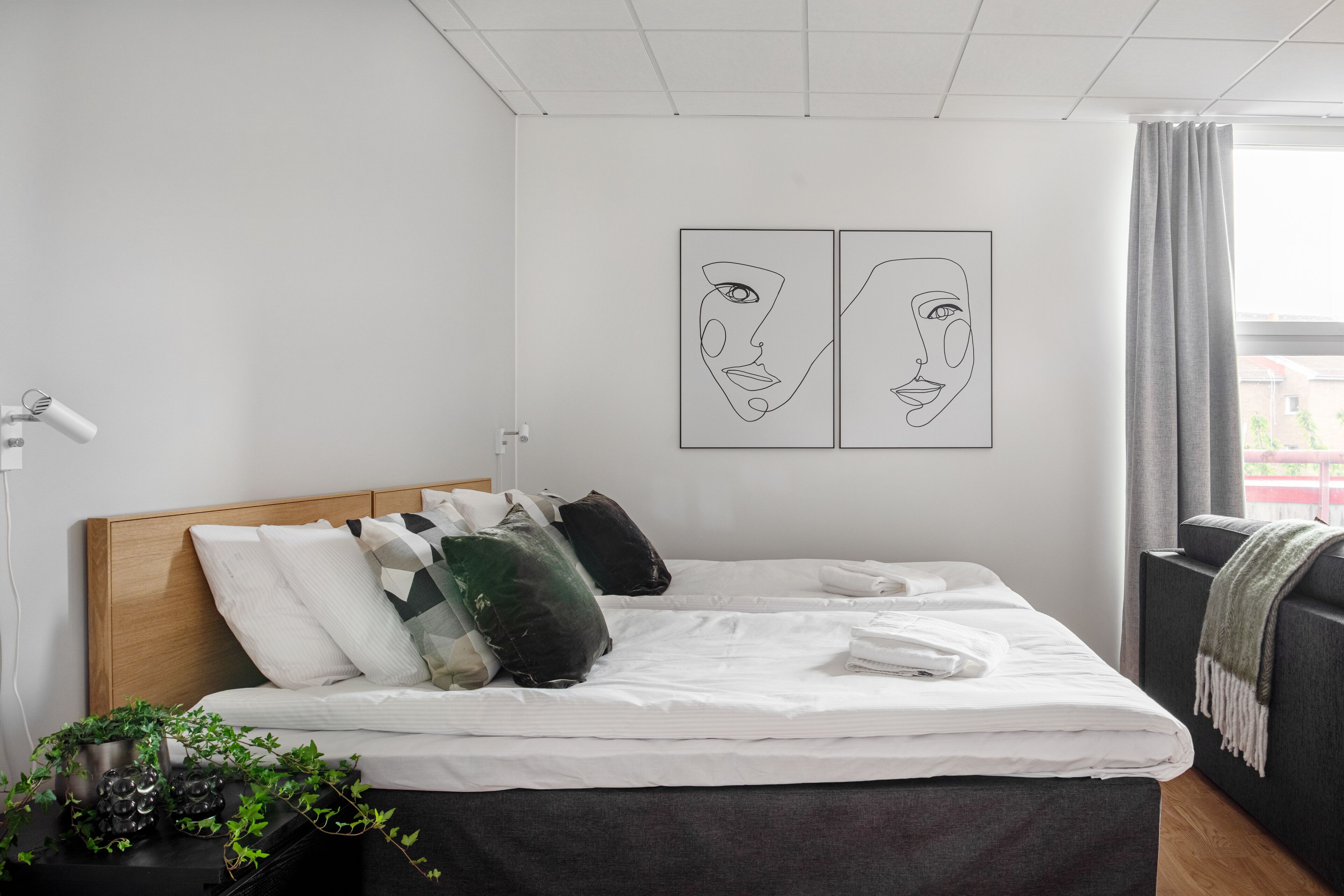 Una cama bien hecha con sábanas blancas, cabecero de madera y varios almohadones. Se observan dos obras de arte colgadas en la pared que representan rostros abstractos. A la izquierda, se ve una mesita con una lámpara y una planta en maceta.