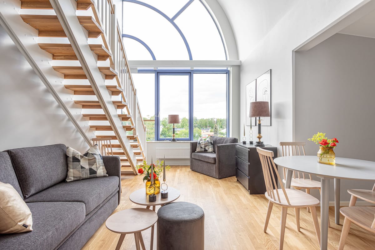 A modern living room with a staircase, a sofa, a dining table, and a view through large windows.