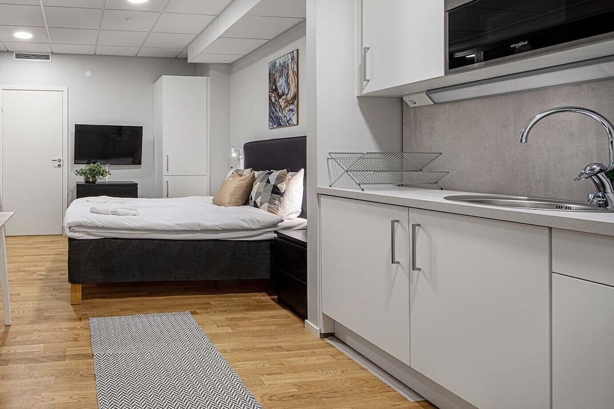 A modern bedroom with a bed, bedside table, and a TV. Adjacent is a kitchenette with a stove, sink, and cabinets.