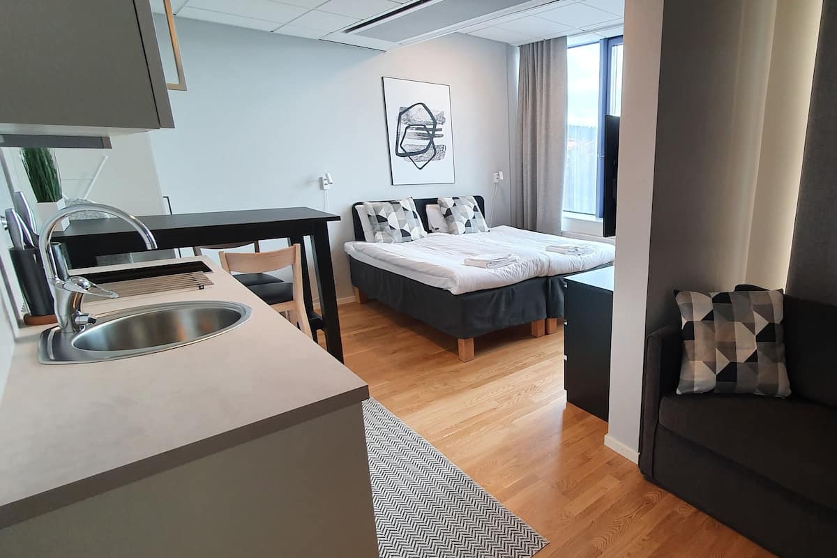 A modern hotel room with a kitchenette, a bed with geometric-patterned pillows, a dark sofa, and a large window with curtains.