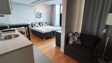 A modern hotel room with a kitchenette, a bed with geometric-patterned pillows, a dark sofa, and a large window with curtains.