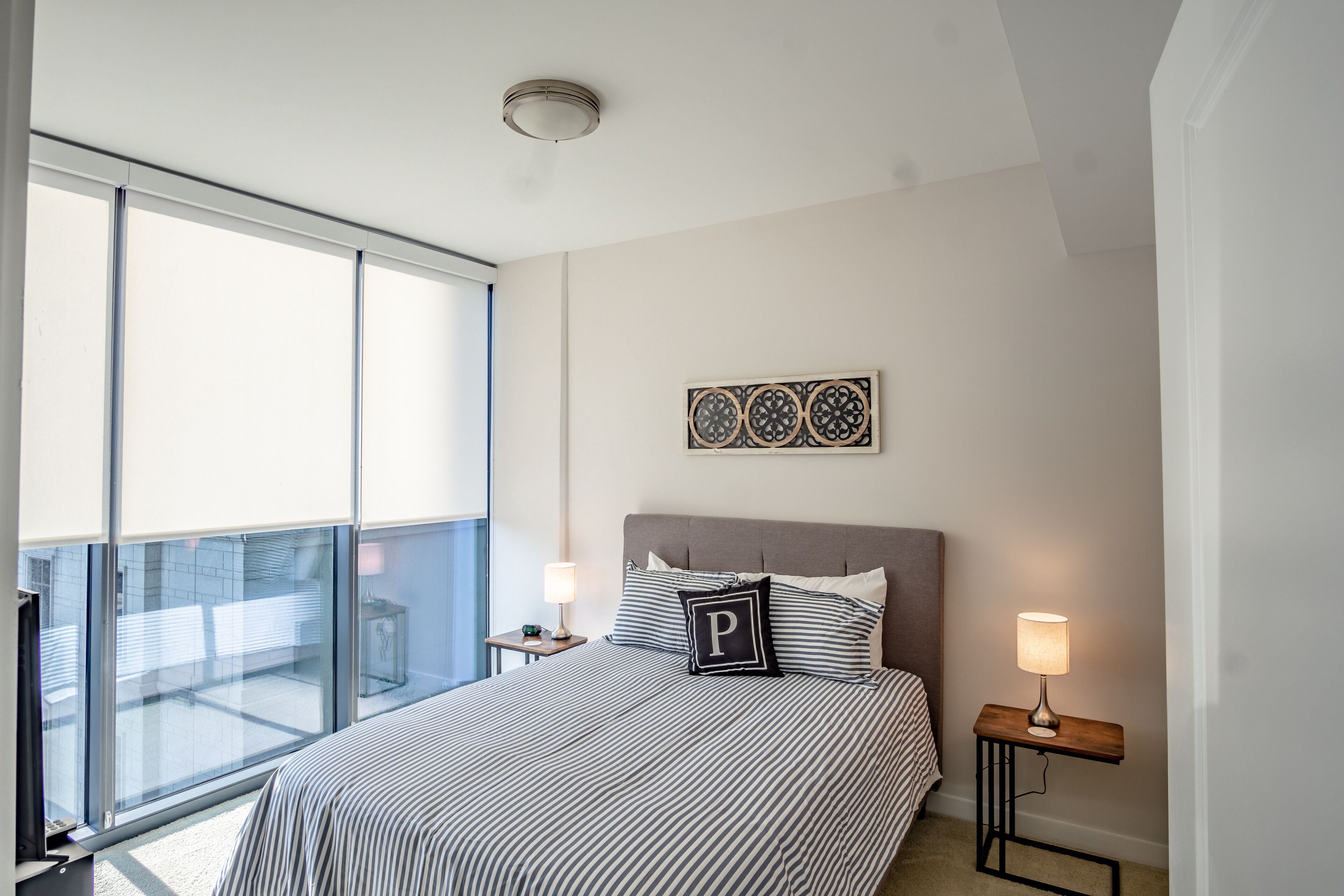 Une chambre avec une grande fenêtre, un lit avec une literie à rayures, une table de chevet avec une lampe et une œuvre d’art accrochée au mur.