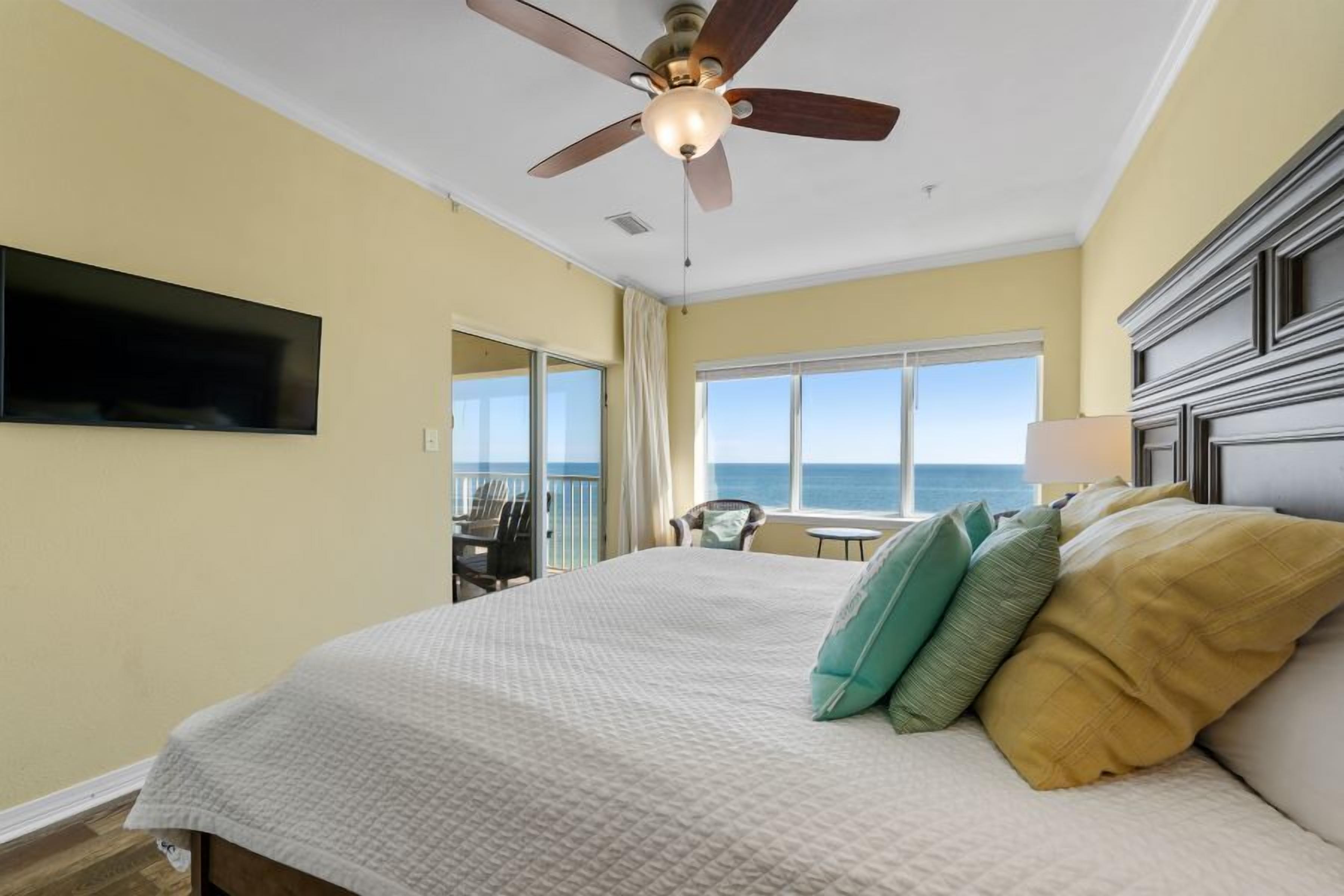 A hotel room with a bed, a ceiling fan, a TV mounted on the wall, and a view of the ocean through large windows.
