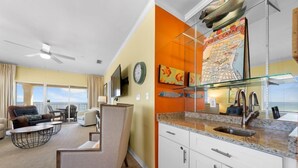 Una sala de estar moderna con sofá, sillón y mesa de centro. Junto a la sala, una cocina con armarios blancos y encimera de granito.