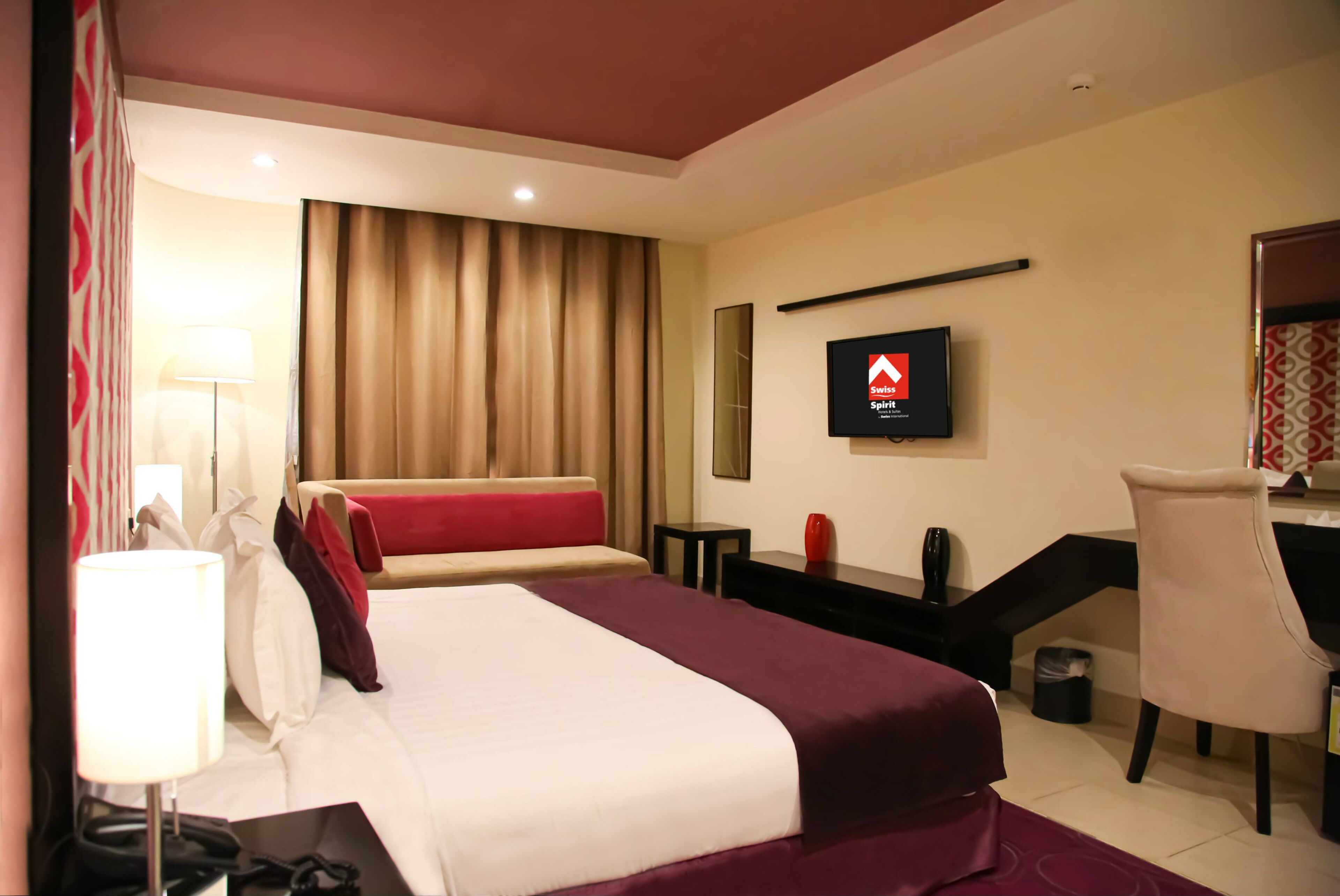 A hotel room with a bed, a desk, a chair, a TV, and a lamp.
