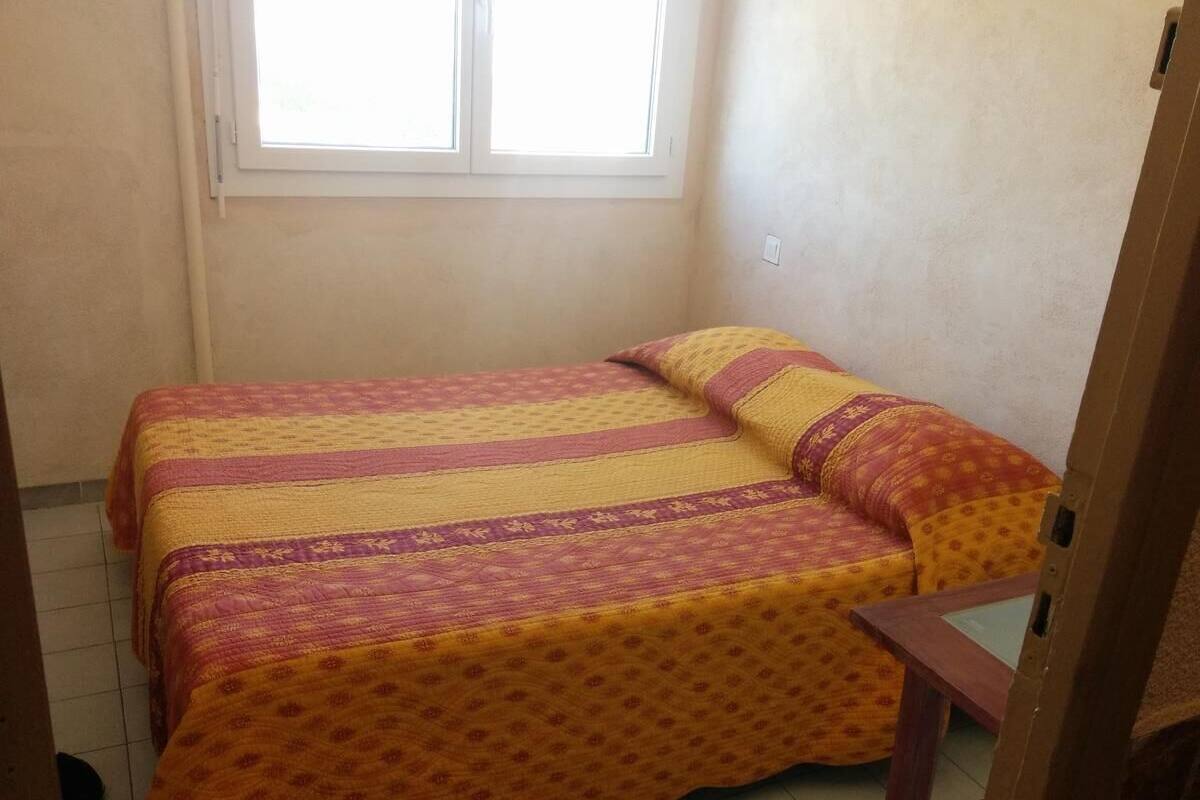 Ein kleines Zimmer mit einem Einzelbett, einer gemusterten Tagesdecke und passenden Kissen. Ein Fenster lässt natürliches Licht herein, und ein Holzhocker steht in der Nähe des Bettes.