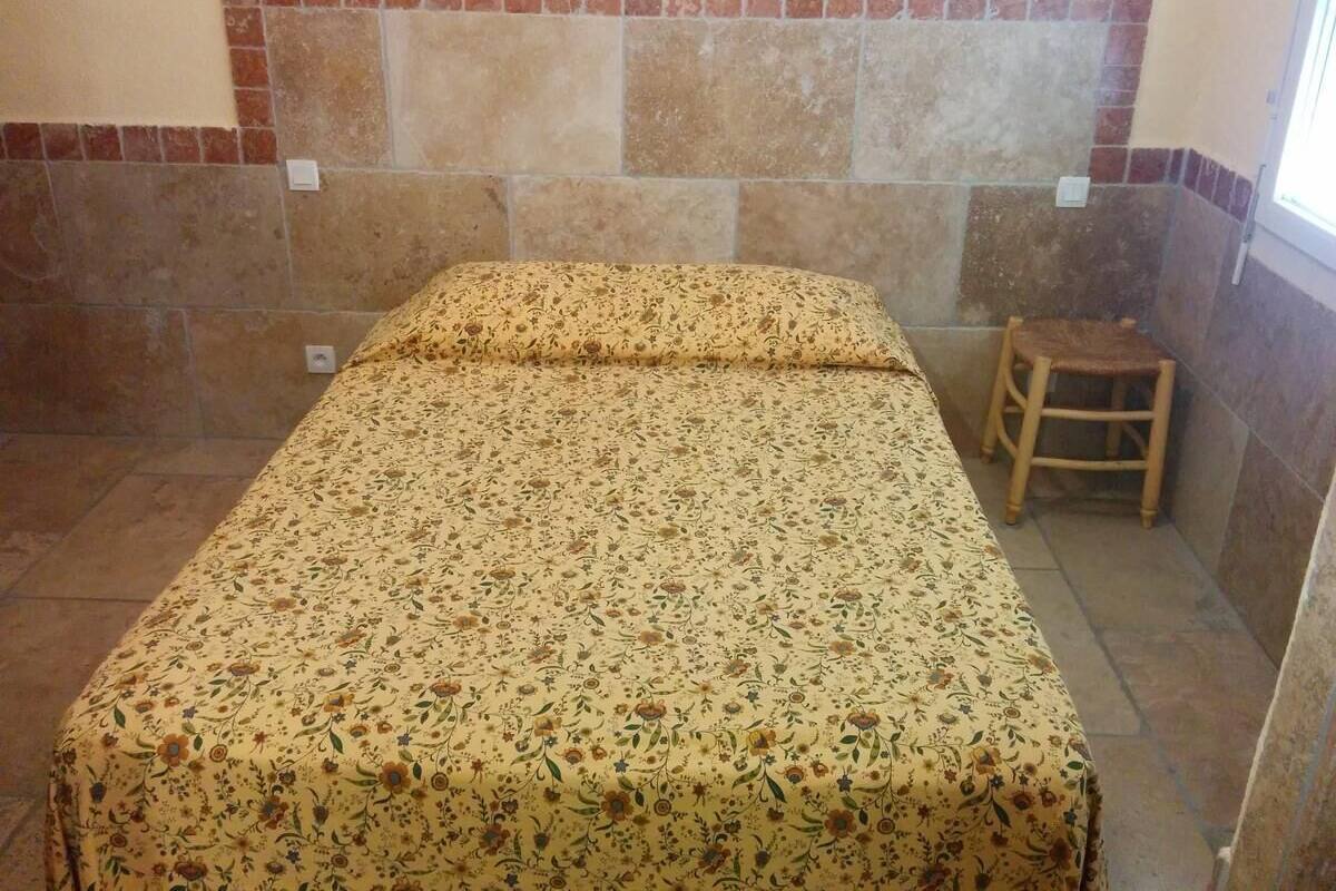 Ein kleiner Raum mit einem Bett, das mit blumigem Bettbezug bezogen ist, einem hölzernen Hocker und einer Wand mit roten Fliesen.