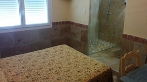 Une salle de bain avec une douche, des toilettes et un lavabo. Il y a une fenêtre avec des stores, un téléviseur fixé au mur et une nappe à motifs sur le sol.