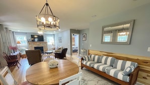 A spacious living area with a wooden dining table, white upholstered chairs, a striped sofa, and a stone fireplace. - Blessed on the New River - Riverfront, WIFI, Small Dog Friendly, Firepit, Gas Grill, Gas Stove (Lansing)