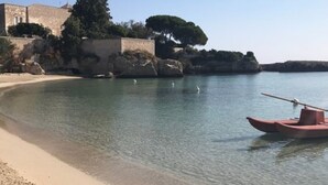 A coastal scene with a clear beach, calm water, a small boat, and a stone building with trees. - La Terrazza dell'Antico Gozzo (Monopoli)