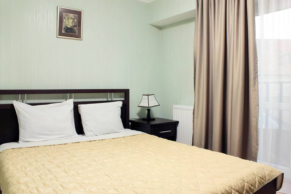 A neatly made bed with white linens and a beige quilt, a bedside table with a lamp, and a framed picture on the wall.