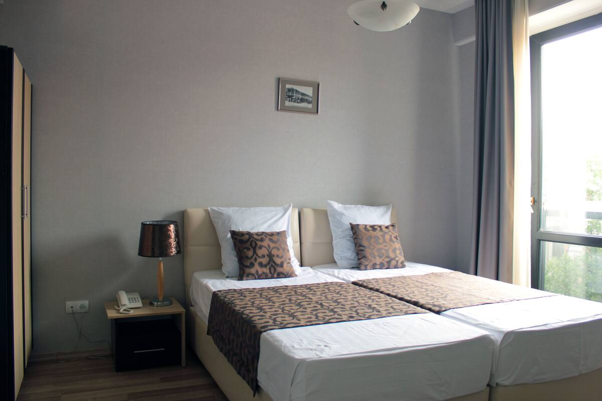 A hotel room with two beds, a bedside table with a lamp, a phone, and a framed picture on the wall.