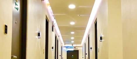 Un largo y estrecho pasillo de hotel con puertas a ambos lados, iluminado por luces en el techo y un cartel direccional verde en la pared izquierda.