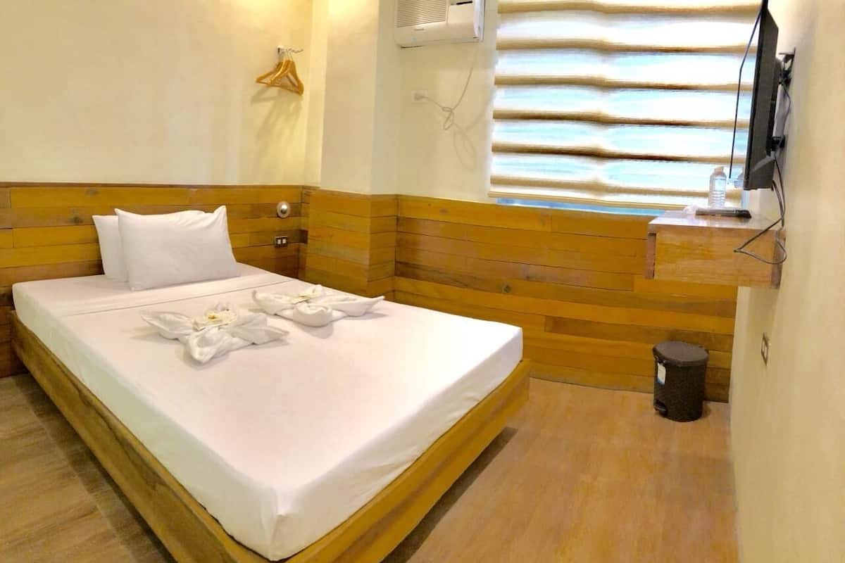 A hotel room with a wooden headboard, a bed with white linens, a small bedside table with a bottle, a wall-mounted TV, and a window with horizontal blinds.