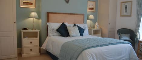 Een keurig opgemaakt slaapkamer met een groot bed, twee nachtkastjes met lampen, een stoel, en lijstjes met foto’s aan de muur.