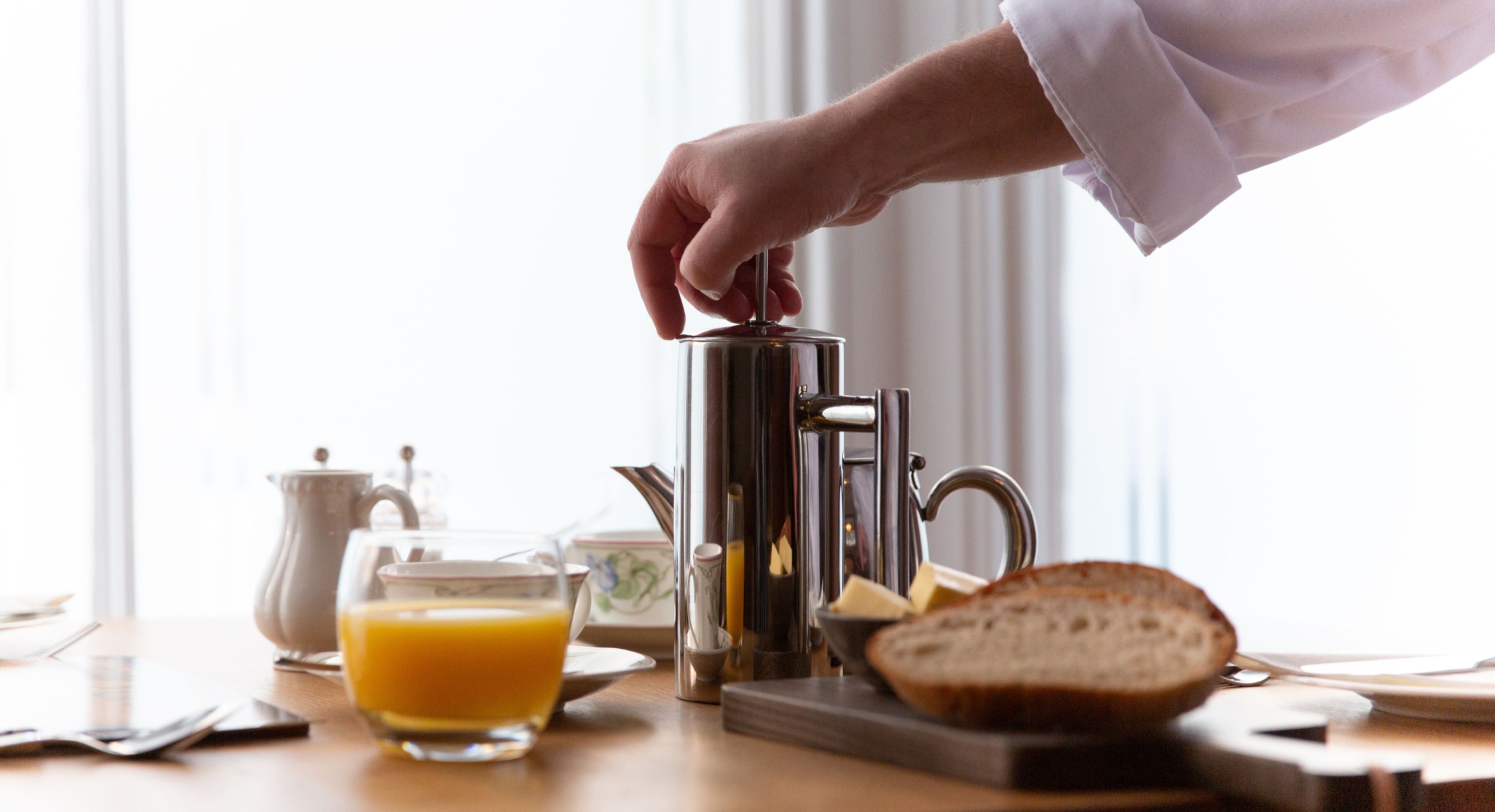 A breakfast setup with a coffee maker, a glass of orange juice, a slice of bread, and a butter dish on a wooden table.