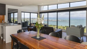 A modern kitchen with a wooden dining table and chairs, a vase with flowers, and large windows overlooking a coastal view. - Wakatu View - Nelson Luxury Holiday Home (Atawhai)