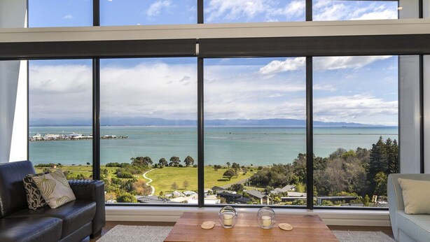 A modern living room with large windows offering a view of a coastal landscape and the sea. - Wakatu View - Nelson Luxury Holiday Home (Atawhai)