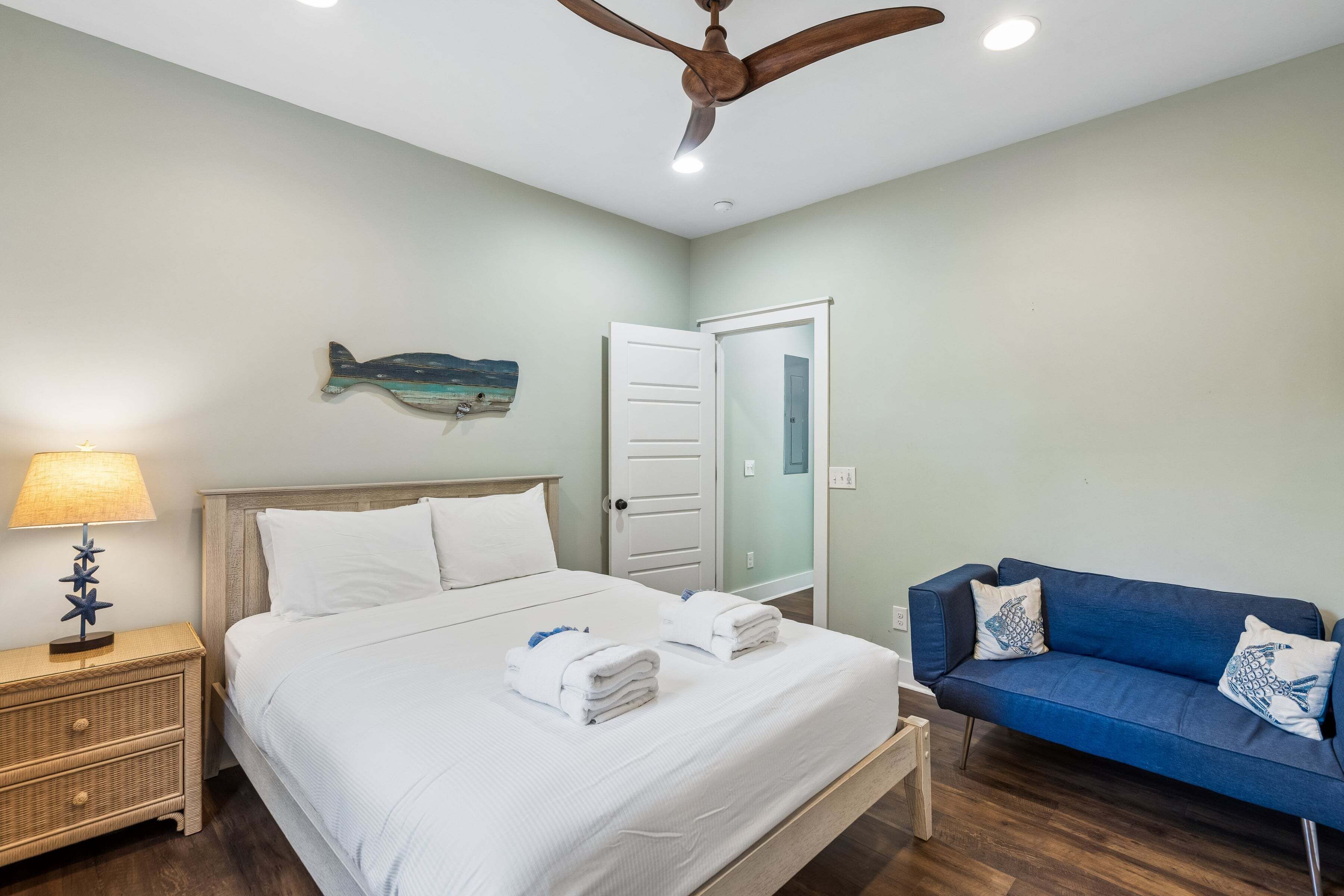 Habitación de hotel con cama, mesita de noche, lámpara, ventilador de techo, sofá azul y una puerta.