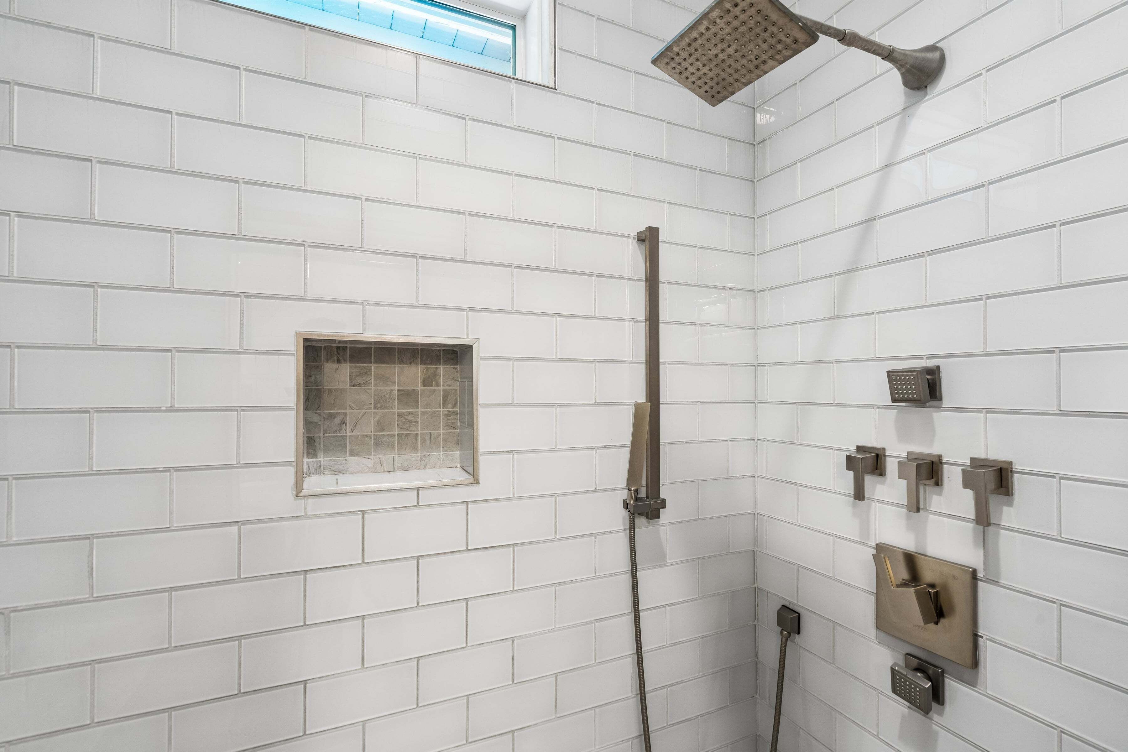 A shower area with white tiled walls, a handheld showerhead, a wall-mounted control panel, and a small window.