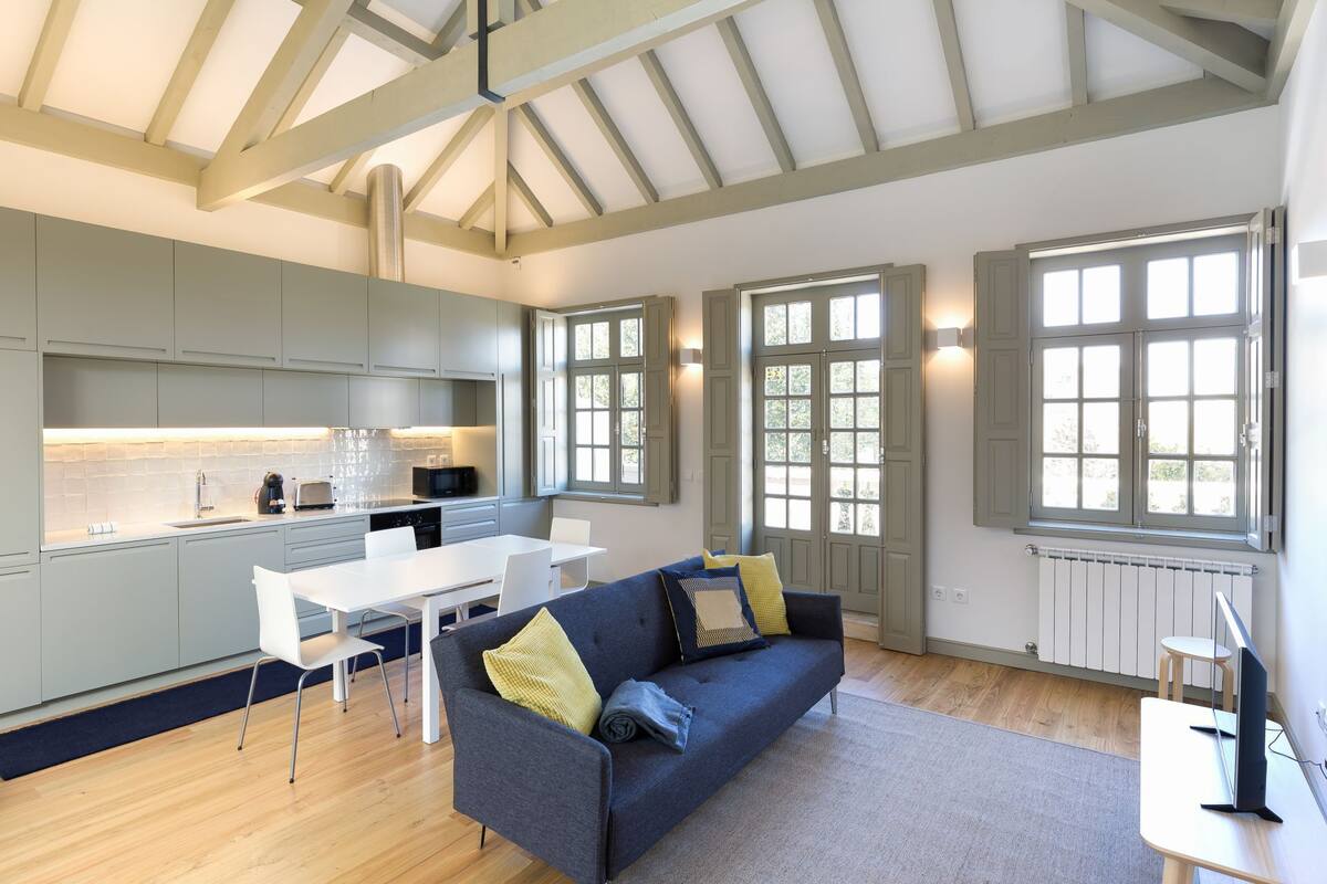 A modern kitchen with grey cabinets, a white countertop, and a dining area with a grey sofa and white chairs.