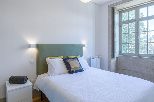 A neatly made bed with white bedding and a green headboard, a bedside table with a rolled-up towel, and a window with a view of trees.