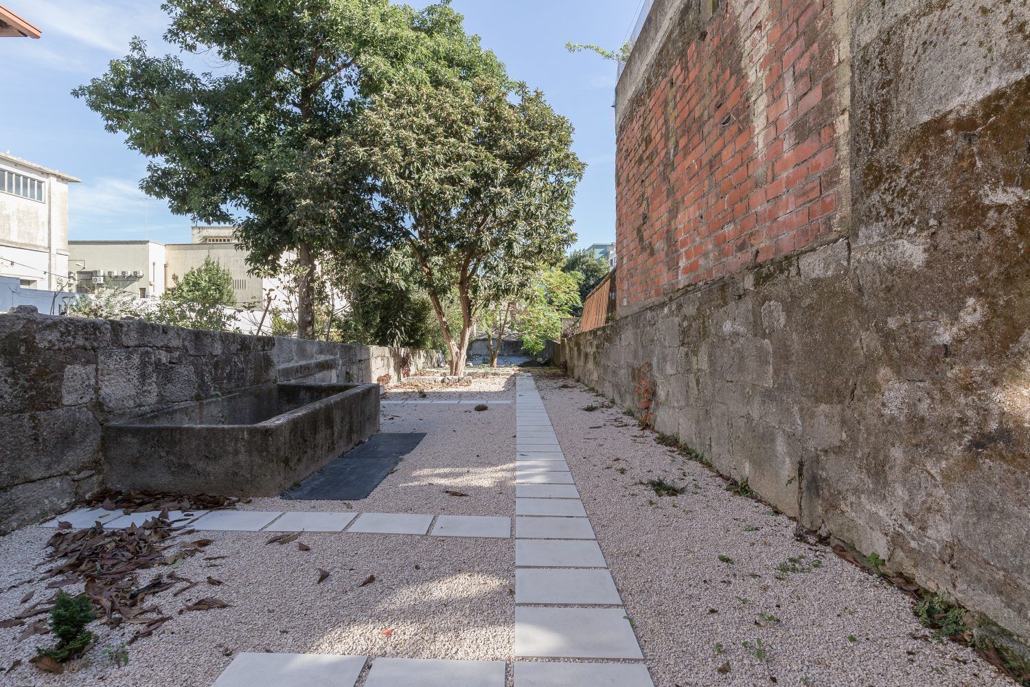 Un sendero estrecho con un banco de piedra, un muro de ladrillo y árboles al fondo.