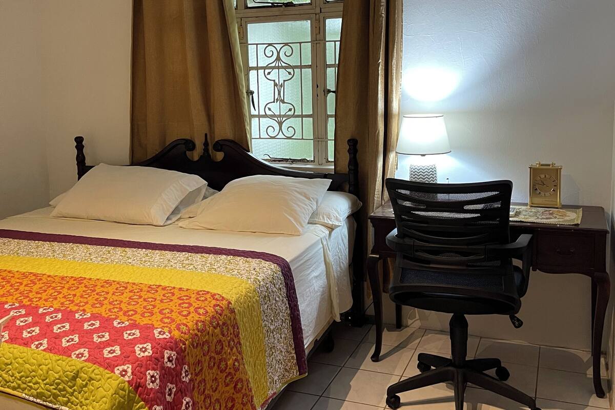 A bedroom with a bed, a desk, a chair, a window with curtains, and a ceiling fan.