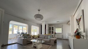 Un salon spacieux avec un canapé beige, une table basse en verre et un meuble blanc. De grandes fenêtres laissent entrer la lumière naturelle et offrent une vue sur l’extérieur.