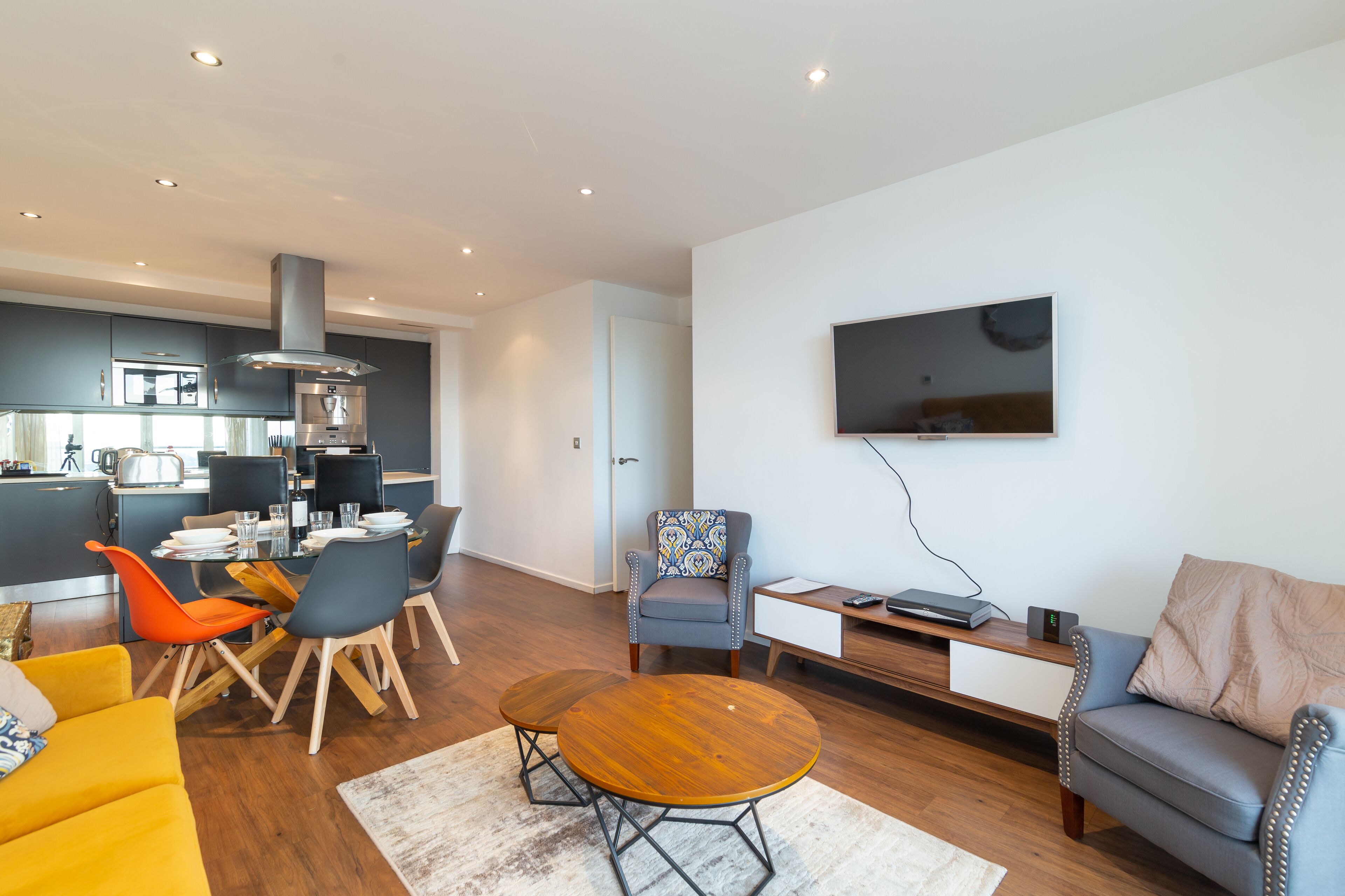 A modern living room with a dining area, a grey sofa, a TV mounted on the wall, and a kitchen in the background.