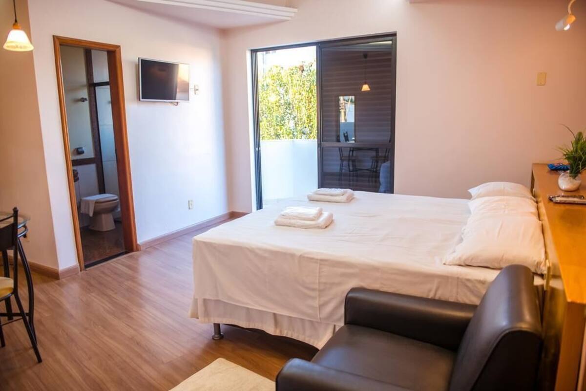 Ein Schlafzimmer mit Bett, Schreibtisch, Stuhl, Fernseher und einem Balkon mit Blick auf Bäume.