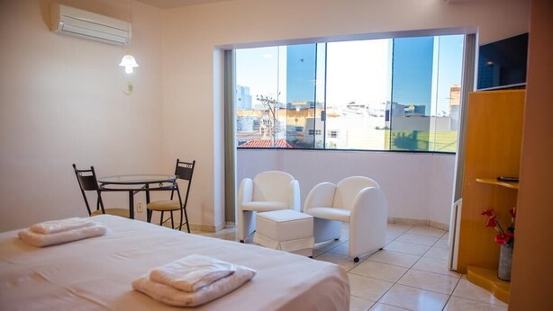 Een hotelkamer met een bed, een kleine tafel met stoelen, een balkon met witte stoelen en een uitzicht op gebouwen door grote ramen.