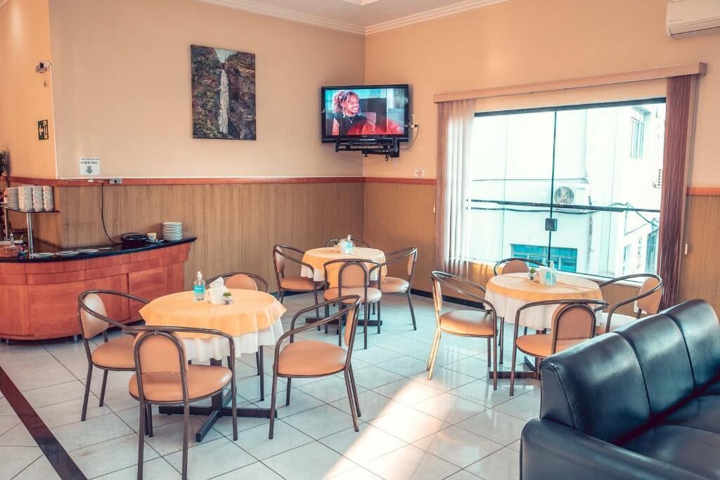 Un vestíbulo de hotel con mostrador de recepción, varias mesas y sillas, un televisor montado en la pared y un ventanal con vistas al exterior.