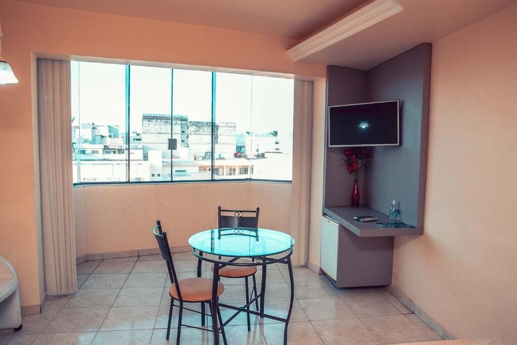 A room with a glass table and chairs, a flat-screen TV, a small shelf, and a large window with a city view.